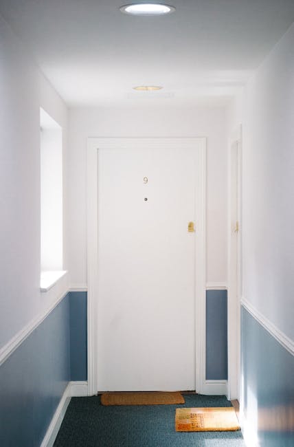 Interior view of a residential hallway leading to a white front door with the number 9, illuminated by soft natural light from two windows on the left. The walls are painted white on the upper portion and feature light blue wainscoting below, separated by a decorative white trim. The flooring consists of a dark, textured carpet, complemented by a brown doormat placed directly in front of the door and a small welcome mat nearby. The clean and well-maintained space exemplifies domestic surface cleaning standards, with no visible dust or clutter, reflecting the professional cleaning services provided by Carpet Cleaning W9 for maintaining hygiene and appearance in flats on Elgin Avenue in Maida Vale W9.